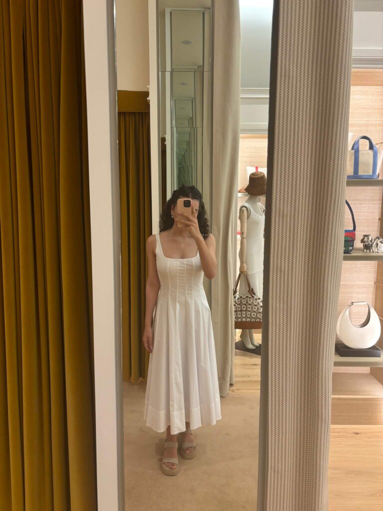 Woman in white STAUD maxi dress standing in dressing room mirror