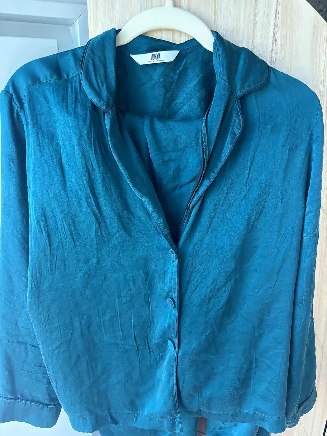 Deep teal Lunya washable silk pajama top with navy piping on a hanger