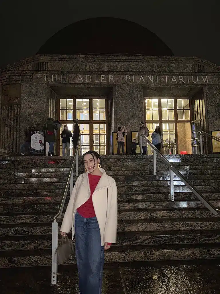 Woman in a cream Marcella virgin wool coat over a red top and jeans, outside the Adler Planetarium at night