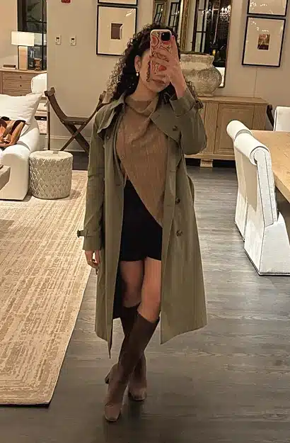 Woman wearing an olive Quince organic cotton trench coat over a tan sweater, black skirt and brown knee-high boots